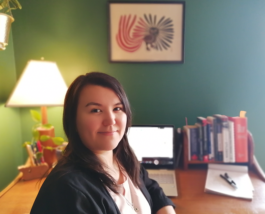 Inuk editor Rachel Taylor sits at a desk. She is smiling at the camera. On the desk is an open laptop and a shelf of books. 